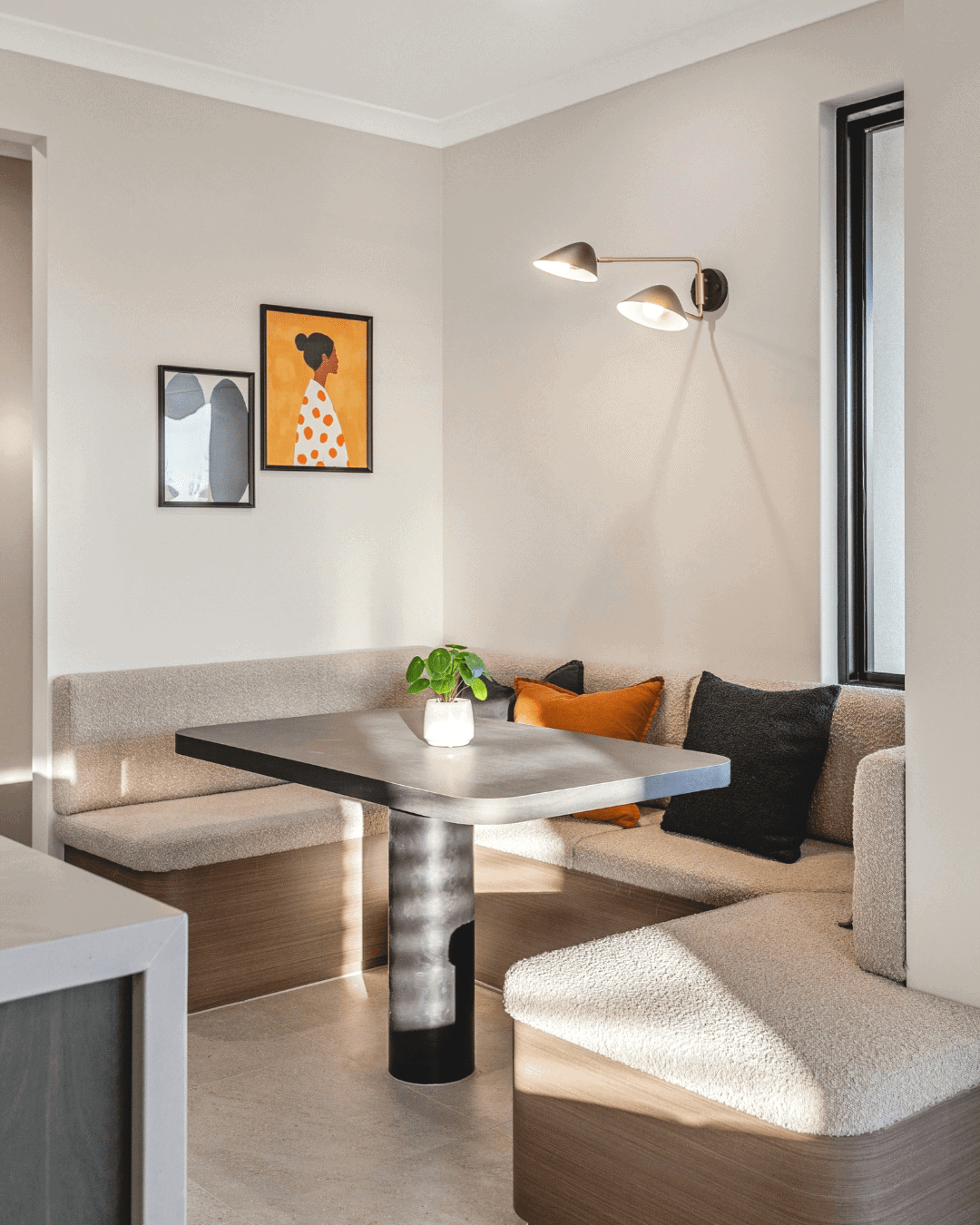 A cozy dining area featuring built-in bench seating, a dark wood table, and decorative cushions, illuminated by natural light and wall-mounted lighting, in a contemporary home.